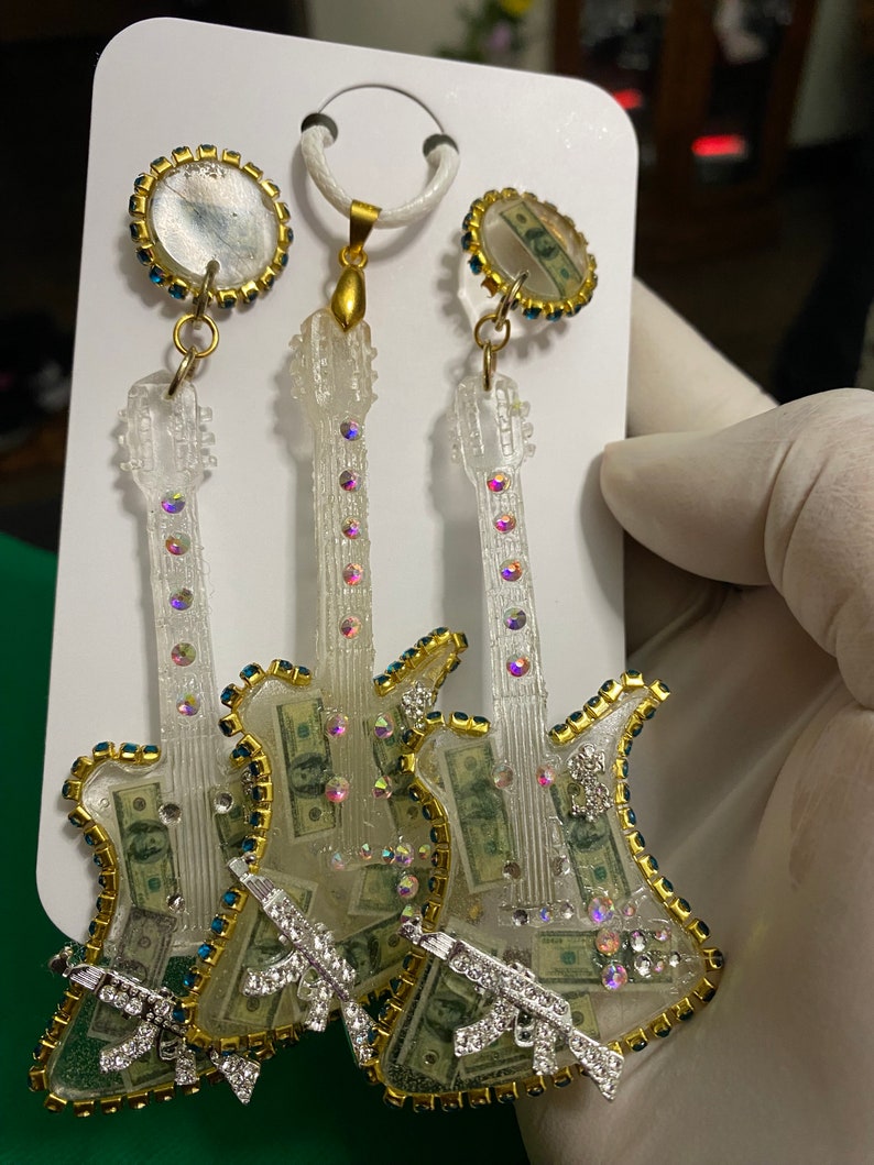 May include: Three clear resin guitar earrings with gold trim and rhinestones. The guitars are filled with images of US dollar bills and have a silver gun charm on each. The earrings are hanging from gold hoops with a clear resin circle with a US dollar bill image.
