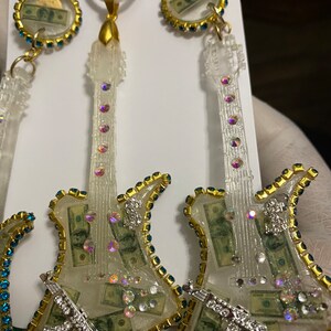 May include: A set of three clear resin guitar-shaped earrings and a pendant necklace. The guitar bodies are filled with dollar bills and rhinestones. The earrings and pendant have gold-colored trim and blue rhinestones around the edges.