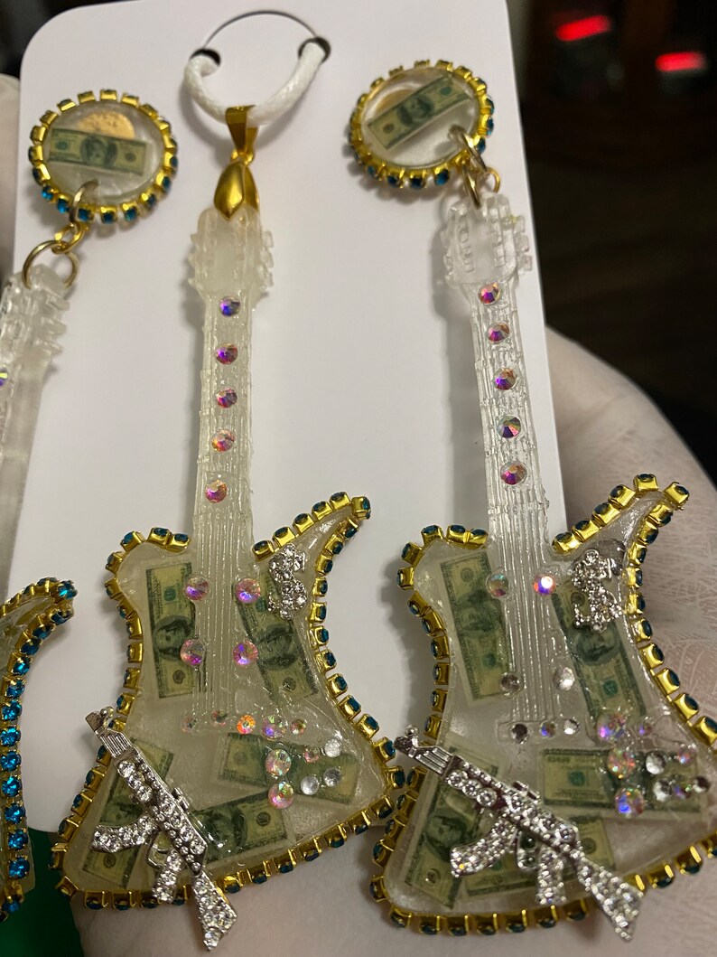 May include: A set of three clear resin guitar-shaped earrings and a pendant necklace. The guitar bodies are filled with dollar bills and rhinestones. The earrings and pendant have gold-colored trim and blue rhinestones around the edges.