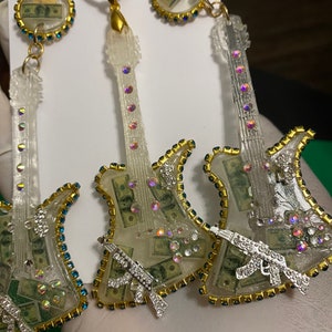 May include: A pair of white guitar-shaped earrings with a gold trim and rhinestones. The guitar bodies are filled with images of US dollar bills and each earring has a silver gun charm.