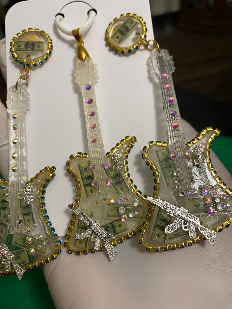 May include: A pair of white guitar-shaped earrings with a gold trim and rhinestones. The guitar bodies are filled with images of US dollar bills and each earring has a silver gun charm.