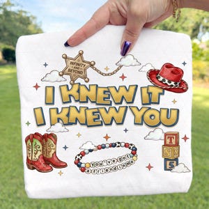 May include: A white t-shirt featuring a graphic print with the text I KNEW IT I KNEW YOU, a sheriff badge with INFINITY AND BEYOND, a red cowboy hat, two red and green cowboy boots, a beaded bracelet with YOU'VE GOT A FRIEND IN ME, and blocks.