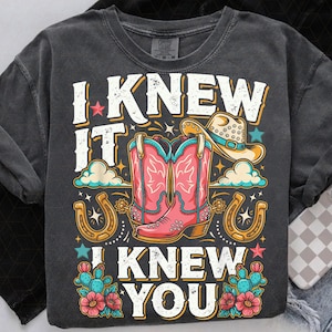 Pu&ograve; includere: Una t-shirt grigio antracite con una stampa grafica che riporta il testo I KNEW IT I KNEW YOU. Il design include un paio di stivali da cowboy rosa, un cappello da cowboy beige, due ferri di cavallo dorati, cactus verde acqua con fiori rosa e piccole stelle.