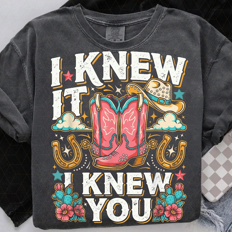 Pu&ograve; includere: Una t-shirt grigio antracite con una stampa grafica che riporta il testo I KNEW IT I KNEW YOU. Il design include un paio di stivali da cowboy rosa, un cappello da cowboy beige, due ferri di cavallo dorati, cactus verde acqua con fiori rosa e piccole stelle.