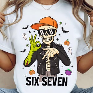 Peut inclure: T-shirt blanc avec un squelette portant des lunettes de soleil et une casquette orange, et une veste en cuir noire. Le squelette fait le signe OK avec une main verte. Le texte "SIX SEVEN" est imprimé en bas. Thème d'Halloween.