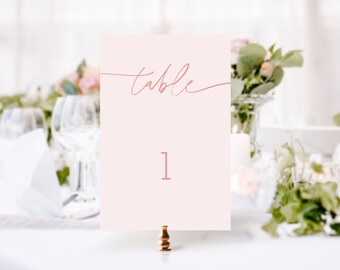 May include: A pink table number card with the word "table" written in cursive script and the number "1" in a larger font. The card is on a gold stand.