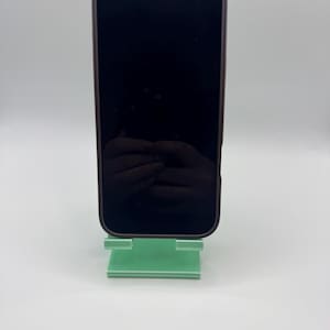 May include: A black smartphone in a dark brown case sits on a mint green phone stand. The phone's screen is dark, reflecting the surroundings. The stand is rectangular with a slot to hold the phone.