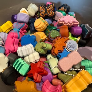 May include: A collection of colorful, small charms in various shapes, including a butterfly, cupcake, star, and cloud. The charms are in shades of pink, yellow, green, blue, and orange. Some charms have small beaded chains attached.