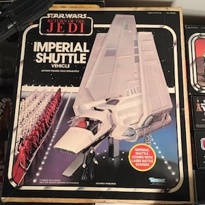 Może przedstawiać: Pudełko z zabawką pojazdem Imperialnego Wahadłowca z filmu Gwiezdne Wojny: Powrót Jedi. Pudełko przedstawia biały i szary Imperialny Wahadłowiec z czerwono-białym paskiem na boku. Pudełko ma również czarne tło z napisem "Star Wars Return of the Jedi Imperial Shuttle Vehicle" w białym kolorze. Pudełko zawiera również napis "Imperial Shuttle comes with laser battle sounds".