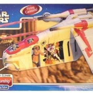 Puede incluir: Un juguete de nave espacial de la República Star Wars blanco y rojo con cañones que disparan. El juguete está en una caja con una imagen de fondo de un paisaje desértico. La caja también presenta el texto "Attack of the Clones" y "Republic Gunship".