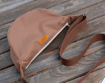 Op de afbeelding: Een bruine canvas crossbody tas met een ritssluiting en een verstelbare bruine schouderband. De tas heeft een klein lederen label met de tekst "HANDMADE" erop.
