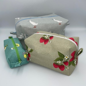May include: Three fabric zipper pouches in varying sizes. The largest is gray with white geese, the medium is tan with red strawberries, and the smallest is teal with a citrus fruit pattern. Each pouch has a contrasting zipper.
