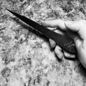 May include: A black letter opener with a sharp, pointed blade. The letter opener has a dark, textured surface and a small, handwritten initial on the side. The object is held in a hand against a patterned background.