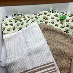 May include: A set of kitchen textiles including two oven mitts with a light green Grogu pattern and two towels. One white towel has brown stripes, and the other is a solid tan color. The oven mitts have large button closures.