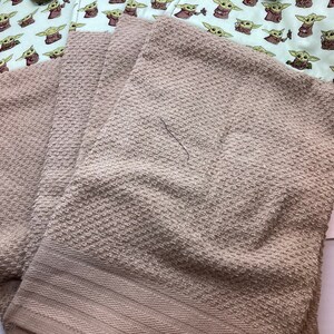 May include: A set of light brown towels with a textured surface, paired with a white and green fabric featuring a repeating pattern of a cartoon character. The fabric has brown buttons and green buttons.