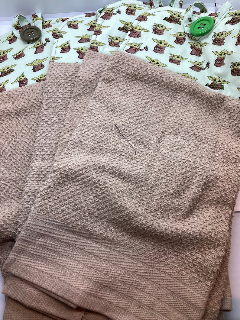 May include: A set of light brown towels with a textured surface, paired with a white and green fabric featuring a repeating pattern of a cartoon character. The fabric has brown buttons and green buttons.