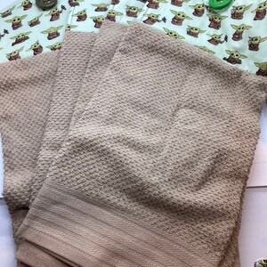 May include: Tan-colored kitchen towels with a textured pattern are stacked below two pot holders. The pot holders feature a light green background with a repeating pattern of a cartoon character. The pot holders have large, decorative buttons.