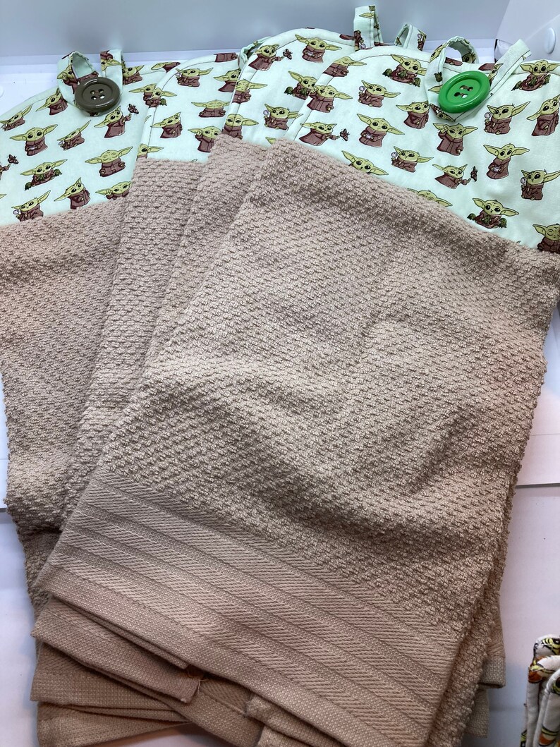 May include: Tan-colored kitchen towels with a textured pattern are stacked below two pot holders. The pot holders feature a light green background with a repeating pattern of a cartoon character. The pot holders have large, decorative buttons.