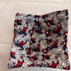 May include: A square, fabric bowl cozy featuring a repeating pattern of the superhero Spider-Man in red and blue, set against a gray background with white spiderwebs. The edges are neatly sewn with a blue thread.