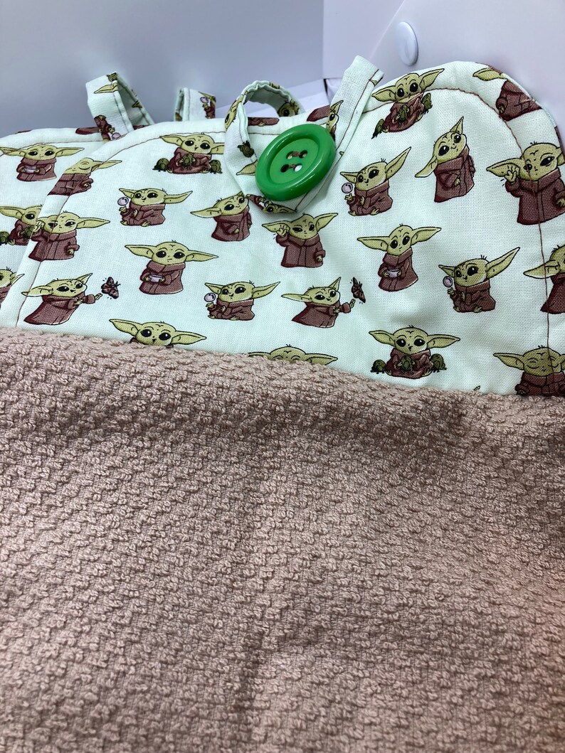May include: A light green and brown towel with a repeating pattern of a cartoon character. The towel has a brown, textured bottom half and a top half with the character print. A large green button is visible.