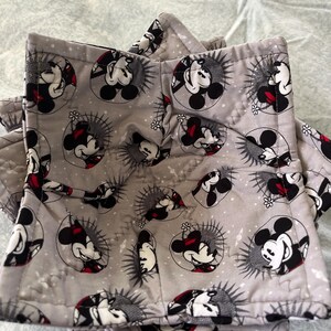 May include: Gray quilted pot holders featuring a repeating pattern of Mickey Mouse in a circular design with red, black, and white accents. The pot holders are square with a folded edge.