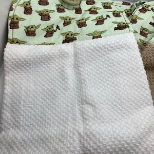 May include: A light green oven mitt and pot holder set featuring a repeating pattern of a cartoon character. The oven mitt has a loop and a large button. A white textured towel is in the foreground.
