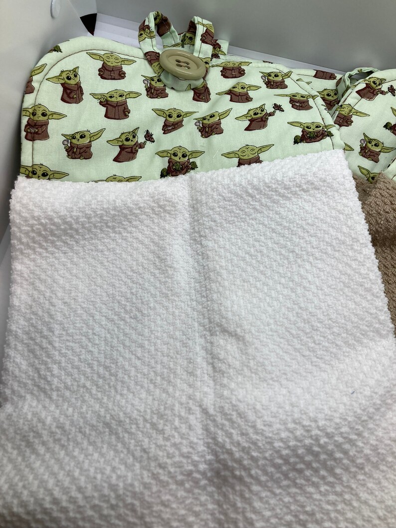 May include: A light green oven mitt and pot holder set featuring a repeating pattern of a cartoon character. The oven mitt has a loop and a large button. A white textured towel is in the foreground.
