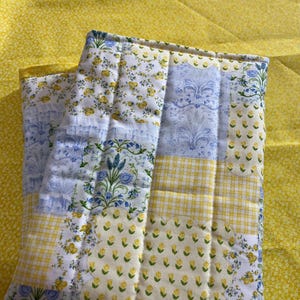 May include: A folded, quilted fabric item with a patchwork design. The quilt features sections of white fabric with yellow floral patterns, blue floral patterns, and yellow gingham. The background is a solid yellow fabric.