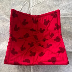 May include: A red, quilted bowl cozy featuring a pattern of dark Mickey Mouse silhouettes. The cozy is open and folded, showcasing the interior lining. The fabric appears soft and padded, designed to insulate and protect.