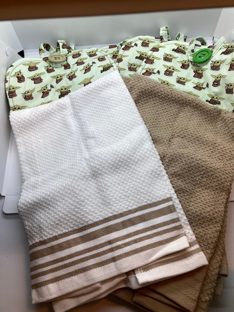 May include: A set of kitchen textiles including two oven mitts with a light green Grogu pattern and two towels. One white towel has brown stripes, and the other is a solid tan color. The oven mitts have large button closures.