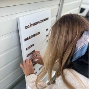 Pode incluir: Uma placa retangular branca com letras marrons, exibindo um cardápio de pizza. O cardápio inclui as palavras "DOŻYNKI PIZZA MENU", "MARGHERITA", "COTTO" e "CRUDO". Uma pessoa está ajustando as letras na placa.