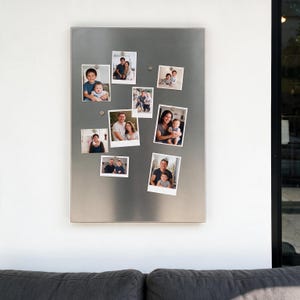 Könnte beinhalten: Eine rechteckige Magnettafel aus gebürstetem silberfarbenem Metall, die an einer Wand befestigt ist. Die Tafel hat eine glatte, metallische Oberfläche und ist mit neun einzelnen quadratischen Fotos von Familien und Kindern verziert, die mit kleinen Magneten befestigt sind.