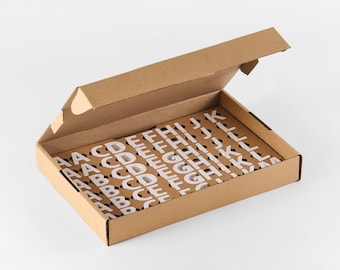 Könnte beinhalten: Ein Satz weißer Magnetbuchstaben, die in einer offenen Pappschachtel angeordnet sind. Die Buchstaben sind in verschiedenen Schriftarten und Größen. Der Text unten lautet "Set of White Magnetic Letters from NOKU".