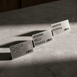Peut inclure: Trois panneaux rectangulaires en métal avec du texte noir sur une surface grise. Les panneaux affichent les mots "Raspberry Danish", "Chocolate Almond Croissant" et "Pistachio Croissant". Les panneaux sont légèrement inclinés.