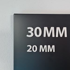 May include: A black sign with the white text "30 MM" and "20 MM". The sign is mounted on a white wall. The text is in a sans-serif font.