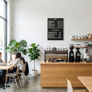 Può includere: Interno di una caffetteria con bancone in legno, macchina per l'espresso e lavagna con menu. I clienti sono seduti ai tavoli. Piante e design minimalista creano un'atmosfera accogliente.