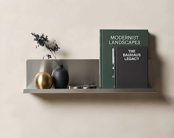 May include: A matte gray metal wall shelf holding a gold sphere, a black vase with a dark branch, a small circular stone coaster, and two books titled Modernist Landscapes and The Bauhaus Legacy with dark green and black covers respectively.