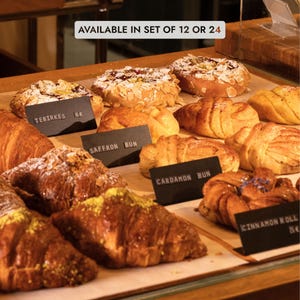 Könnte beinhalten: Eine Auswahl an Backwaren, darunter Croissants und Brötchen, auf einer Holzoberfläche. Die Backwaren sind goldbraun und mit Puderzucker und Nüssen bestreut. Schwarze Schilder kennzeichnen die Namen der Backwaren. Der Text oben lautet "AVAILABLE IN SET OF 12 OR 24."