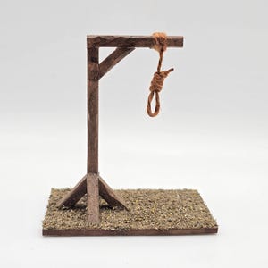 May include: A miniature gallows constructed from brown wood, with a noose suspended from the crossbeam. The base is a square of brown wood covered in dried plant matter. The text "Nietzsche Miniatures" is visible at the top.