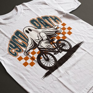 Peut inclure: T-shirt blanc avec un dessin d'un fant&ocirc;me de dessin anim&eacute; &agrave; v&eacute;lo. Le fant&ocirc;me est blanc avec des yeux noirs, faisant du v&eacute;lo noir. Le texte "CYCO OATH" est au-dessus du fant&ocirc;me en bleu sarcelle et orange. Un motif &agrave; carreaux est en arri&egrave;re-plan.
