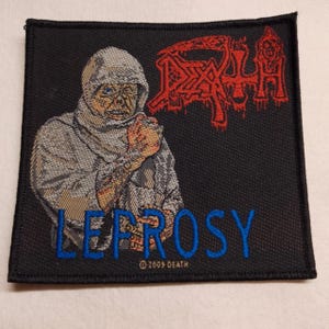 May include: A square black embroidered patch featuring a hooded figure in grey tones. The red text reads DEATH, and the blue text reads LEPROSY. Below the blue text, the small white text reads 2009 DEATH. The patch has a black stitched border.