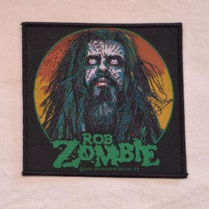 May include: A square embroidered patch featuring a portrait of a person with long dark hair and a beard against a circular orange and yellow background. The text ROB ZOMBIE appears in green below the portrait, with 2012 SPOOKSHOW DELUXE LTD below.