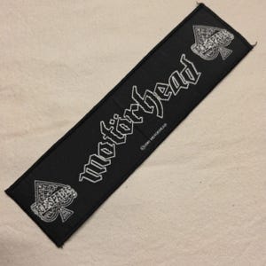 Può includere: Una toppa rettangolare nera ricamata con il testo bianco Motorhead in un carattere stilizzato. Il design include un simbolo dell'Asso di picche nell'angolo in alto a destra e in basso a sinistra, con il testo 2010 MOTORHEAD in piccole lettere bianche.