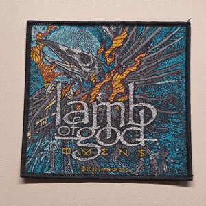 Könnte beinhalten: Ein quadratischer gestickter Aufnäher mit einer stilisierten Vogelschädelgrafik und orangefarbenen Flammen auf türkisfarbenem und schwarzem Hintergrund. Der Text lautet lamb of god, Omens und 2022 LAMB OF GOD. Der Aufnäher hat einen dicken schwarzen Rand.
