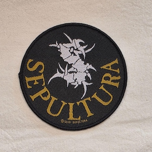 Puede incluir: Un parche bordado circular de color negro con un gráfico tribal blanco estilizado en el centro. La palabra SEPULTURA está bordada con hilo dorado a lo largo de la curva inferior. El texto 2010 SEPULTURA es visible en letras doradas pequeñas.