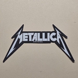 Puede incluir: Un parche bordado en blanco y negro que presenta el logotipo de Metallica con letras angulares y afiladas. El diseño incluye el texto Metallica en una fuente estilizada y el pequeño texto de copyright 2013 Metallica centrado bajo la palabra.