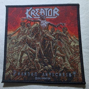 May include: A square woven fabric patch featuring the Kreator logo at the top and the text Phantom Antichrist at the bottom. The design depicts a central figure on horseback surrounded by multiple figures against a red and orange background.
