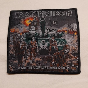 May include: A square woven patch featuring the Iron Maiden logo at the top and the text A MATTER OF LIFE AND DEATH at the bottom. The graphic depicts a military tank surrounded by skeleton soldiers in a battlefield scene with orange explosions.