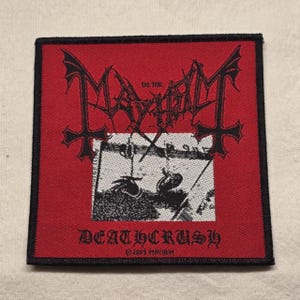 Könnte beinhalten: Ein quadratischer Stoffaufnäher mit rotem Hintergrund und schwarzem Rand. Er zeigt den schwarzen Schriftzug "The True Mayhem" in stilisierter Schrift über einer schwarz-weißen Grafik einer Hand, mit "DEATHCRUSH" und "©2005 MAYHEM" darunter.