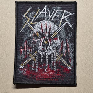 May include: A rectangular embroidered patch featuring a white skull with red markings, two crossed swords, and hanging chains against a dark background. The word SLAYER is stitched in white at the top, with the text 2020 SLAYER printed at the bottom.